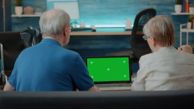 Senior couple using laptop with green screen in living room. Elder man and woman looking at blank chroma key with isolated copy space background and mock up template on computer display.