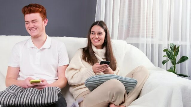 Young beautiful couple sitting on the sofa at home in casual clothing holding mobile phones in their hands and looking at them while somebody threw a paper plane to them.