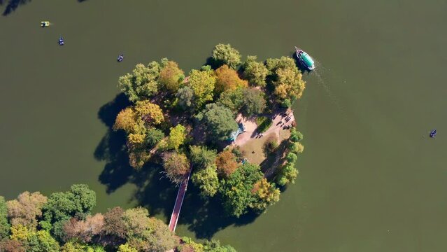Top view of the lake and the island. People swim on a boat, boats on the lake. Yellow, green, autumn leaves, trees from above. People are walking in the park.