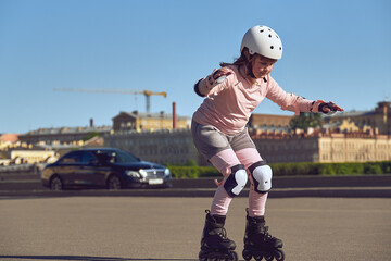 A girl in protection is rollerblading in the city. The child learns to roller skate.