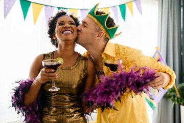 Cheerful black woman and Caucasian man drink cocktails while celebrating Mardi Gras at home party.
