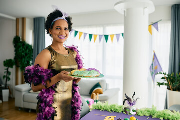 Happy African American woman in Mardi Gras costume serving King cake at home party.