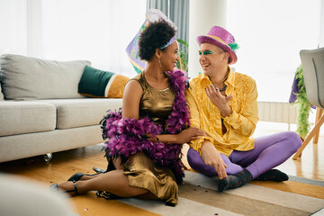 Happy Black woman and her Caucasian friend wear Mardi Gras carnival costumes and talk at home party.