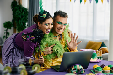 Happy couple in carnival costumes making video call over laptop during Mardi Gras celebration.