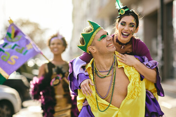 Playful couple have fun while piggybacking on Mardi Gras street parade during the festival. - obrazy, fototapety, plakaty
