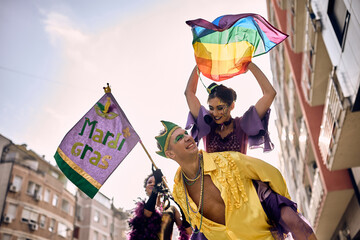 Happy couple with rainbow flag piggybacking on street carnival during Mardi Gras celebration.