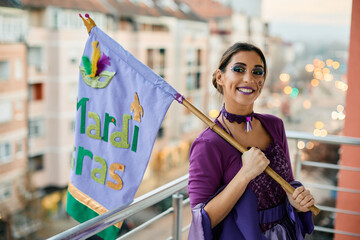 Young woman in carnival costume and make-up standing with Mardi Gras flag on balcony.