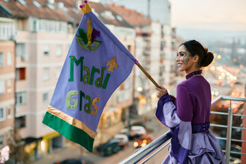 Young woman with Mardi Gras flag standing on balcony during celebration carnival. © Drazen