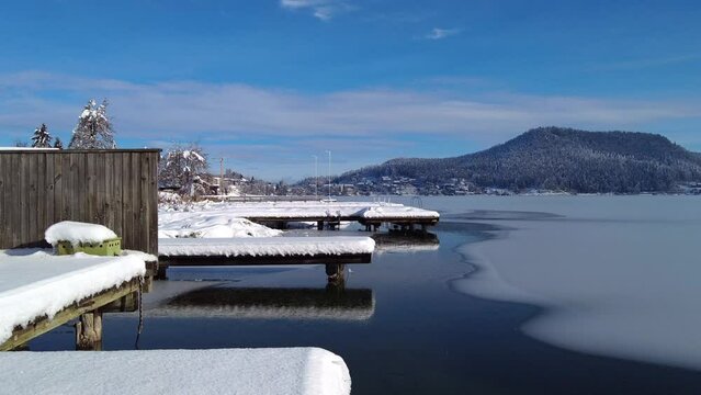 A panoramic view on the Faaker lake in Austrian Alps. The lake is surrounded by high mountains. There is a snow covered pier going into the frozen lake.