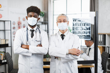 Young african american and aged caucasian doctors in face masks and lab coats posing on camera indoors. Two medical specialists standing near flipchart and holding arms crossed.