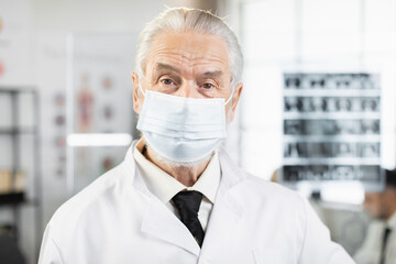 Qualified aged doctor in face mask, lab coat and eyeglasses standing at hospital room and looking at camera. Caucasian medical worker posing during working process.