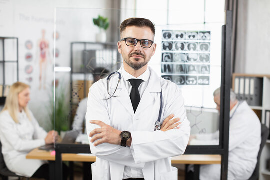 Handsome caucasian man in eyeglasses and lab coat posing with crossed arms at conference room. Medical specialist with stethoscope on neck standing indoors.