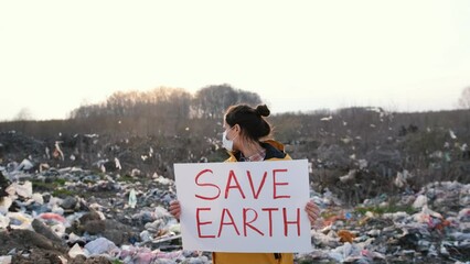 A young woman activist with a poster in her hands saves the planet stands in the middle of a landfill wearing a mask. Female student protests against environmental pollution, nature conservation