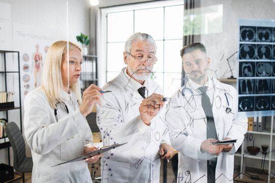 Group of three male and female medical scientists in white lab coats writing on flipchart while examining results of tomography. Competent doctors having meeting at hospital office.