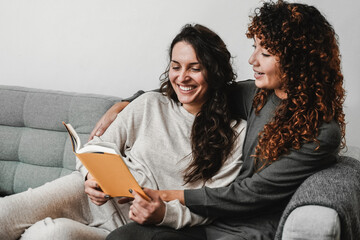 LGBT Hispanic lesbian couple reading a book wearing pajamas in sofa at home - Gay love relationships concept - Focus on left woman face - obrazy, fototapety, plakaty