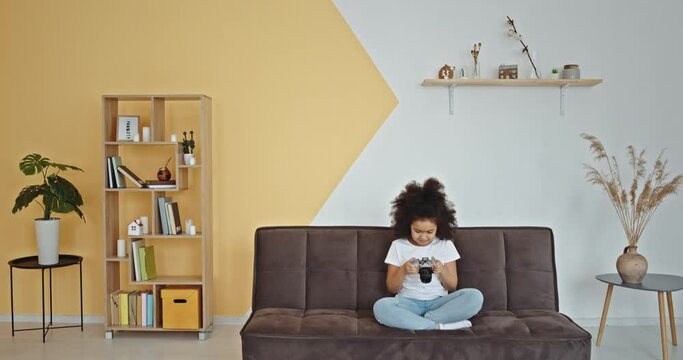 Ethnic girl taking photos on sofa. Mixed race girl with curly hair taking and checking photos with camera while sitting cross legged on couch in living room at home