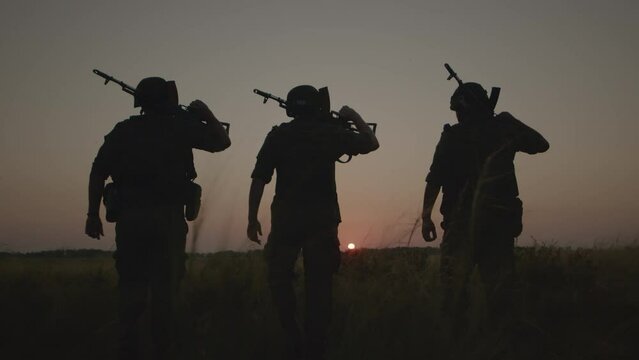 Silhouettes of three fully equipped armed soldiers walking across battle field.