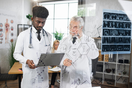 Caucasian aged doctor and his african american assistant standing with modern laptop near glass flipchart. Two scientists examining formula of new virus at hospital room.
