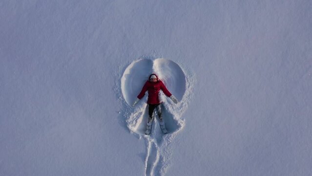 Aerial view from drone of girl in red winter coat making snow angel. Girl waves arms and legs lying in snow. High quality 4k footage