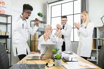 Group of multicultural doctors smiling and clapping in hands while looking on laptop screen during...