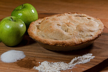 Fresh baked double crust apple pie displayed with key ingredients of green tart apples, sugar, cinnamon, and flour. Other ingredients include vegetable shortening, butter and water to make the dough.