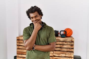 African man with curly hair wearing sportswear at the gym looking confident at the camera with smile with crossed arms and hand raised on chin. thinking positive.