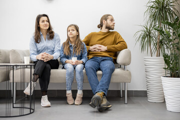 Young family with ten year old girl sitting on a couch at the waiting room of the clinic. Patients are waiting for a doctor's appointment at reception