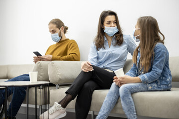 Young woman with a girl and adult man sitting on a couch at the waiting room of the clinic. Patients in face mask waiting for a doctor's appointment at reception