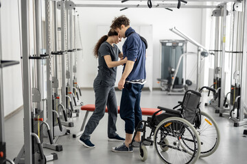 Rehabilitation specialist helps a guy stand out of a wheelchair at rehabilitation center. Concept of physical therapy and support for people with disabilities - obrazy, fototapety, plakaty
