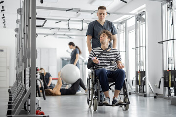 People exercising at rehabilitation center, rehabilitologist walking with guy in a wheelchair. Concept of kinesiology and recovery from injuries