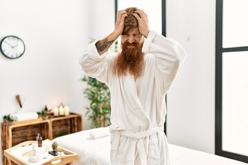 Redhead man with long beard wearing bathrobe at wellness spa suffering from headache desperate and stressed because pain and migraine. hands on head.