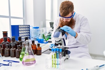 Young redhead man wearing scientist uniform using microscope at laboratory