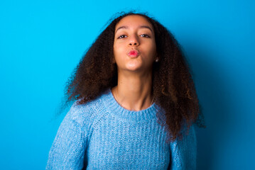 African teenager girl wearing blue sweater over blue background, keeps lips as going to kiss someone, has glad expression, grimace face. Standing indoors. Beauty concept.