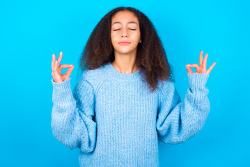African teenager girl wearing blue sweater over blue background doing yoga, keeping eyes closed,...