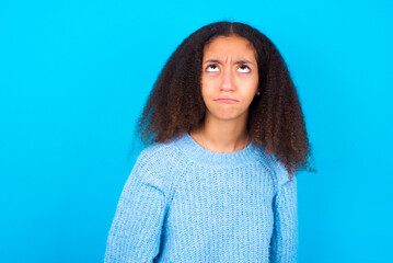 Fototapeta premium African teenager girl wearing blue sweater over blue background has worried face looking up lips together, being upset thinking about something important, keeps hands down.