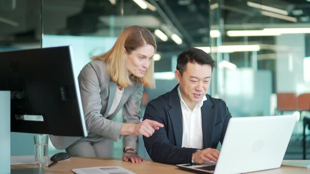 happy coworkers discuss project strategy by looking and pointing at laptop pc computer monitor screen. multiethnic business team in the office. confident mature asian man explaining young female