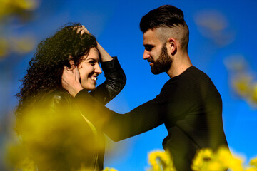 portrait of a young smiling couple where hair man caresses his girlfriend's face before kissing blue background and yellow flowers out of focus in the foreground