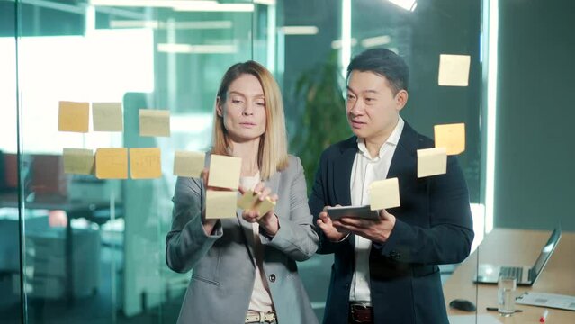 Startup and implementation of a small business project. Asian Businessman discussing with businesswoman sticking adhesive notes on glass wall in office, discussing ideas on visualization board