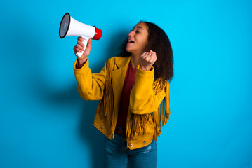 African teenager girl wearing yellow jacket over blue background communicates shouting loud holding a megaphone, expressing success and positive concept, idea for marketing or sales.