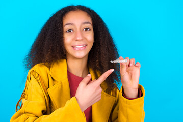 African teenager girl wearing yellow jacket over blue background holding an invisible aligner and...