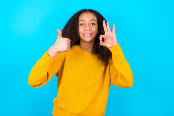 African teenager girl wearing yellow sweater over blue background smiling and looking happy, carefree and positive, gesturing victory or peace with one hand