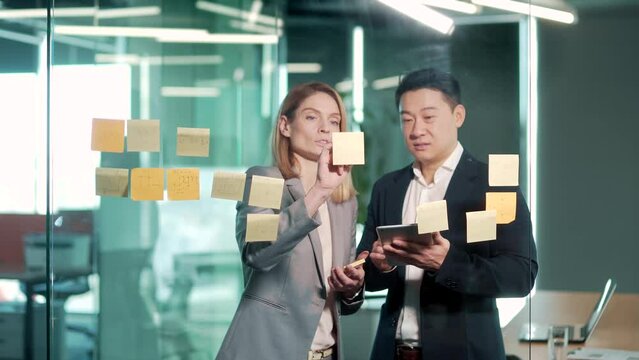 Teamwork meeting in office. Two happy business Asian colleagues team write on post it sticky notes look at glass wall scrum board brainstorm on work organization task management discuss strategy plan
