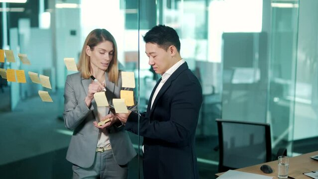 Startup and implementation of a small business project. Asian Businessman discussing with businesswoman sticking adhesive notes on glass wall in office, discussing ideas on visualization board 