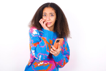 Afraid funny African teenager girl wearing colorful sweater over white background holding telephone and bitting nails