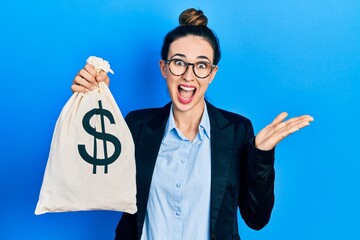 Young hispanic girl wearing business clothes holding dollars bag celebrating achievement with happy...