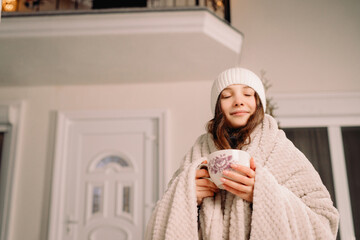 Pretty young girl in white hat and blanket with a cup of tea in her hands closed eyes standing near the house on evening winter holiday.