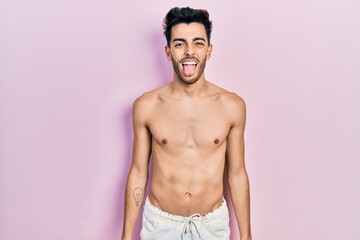 Young hispanic man wearing swimwear shirtless sticking tongue out happy with funny expression....