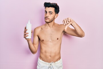 Fototapeta premium Young hispanic man wearing swimsuit and holding sunscreen lotion with angry face, negative sign showing dislike with thumbs down, rejection concept