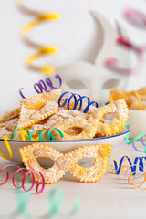 Angel wings or sfrappole or chiacchiere in shape of carnival mask. Traditional sweet crisp pastry deep-fried and sprinkled with sugar. Carnival food. Decorated with paper serpentine. Vertical image. - obrazy, fototapety, plakaty