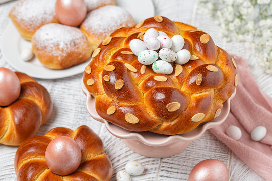 Easter table with italian style homemade braid buns, greek style tsoureki bread with almond, pink metallic colored eggs and festive candies. White flowers. High angle view.